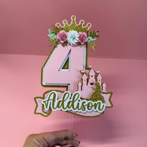 May include: Pink and gold glitter "4" cake topper with a crown, flowers, and a castle design. The name "Addison" is written on a banner. The topper is on a clear stick, perfect for a fourth birthday celebration.