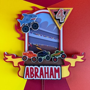 May include: A colorful birthday cake topper featuring a shark on a monster truck, a number 4, and the name "ABRAHAM". The design includes three additional monster trucks in orange, black, and green. The background is a race track scene.