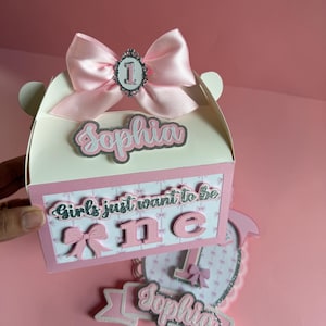May include: A pink and white birthday gift box with a large pink bow and the name "Sophia". The box has the text "Girls just want to be ONE" and a decorative "1". A matching cake topper is also visible.