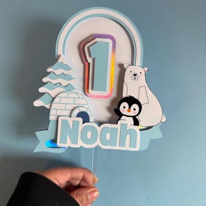 May include: A light blue and white birthday cake topper with a winter theme. The topper features a number 1, a polar bear, a penguin, an igloo, and a Christmas tree. The name "Noah" is also included.