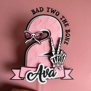 May include: A pink and black birthday cake topper with the number 2, the text "BAD TWO THE BONE", and the name "Ava". The number 2 has a bone pattern and a skeleton hand making a peace sign.