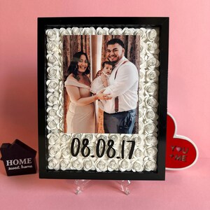 May include: A black picture frame with a white paper flower border. The frame contains a photo of a family of three. The date 08.08.17 is written in black on the bottom of the frame.