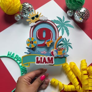 May include: A layered cake topper featuring a large red number nine, a yellow sun with sunglasses, a blue pool float, a water gun, and a beach ball. A teal banner at the bottom displays the name LIAM in white letters. The design includes palm tree silhouettes.