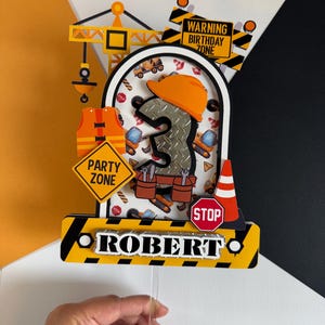 May include: A construction-themed birthday cake topper with the name "ROBERT". The topper features a number 3, an orange hard hat, a crane, and construction vehicles. It includes signs that say "Party Zone", "Warning Birthday Zone", and "Stop".