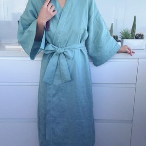 May include: A light teal-colored linen robe with a matching tie belt. The robe has wide sleeves and a relaxed fit. The model is wearing the robe indoors. The robe is knee-length and appears to be made of a soft, breathable fabric.