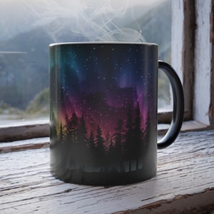 May include: Black ceramic mug featuring a colourful aurora borealis and silhouette of trees design. Steam rises from the mug, suggesting a hot drink.