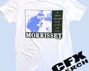Può includere: Maglietta bianca con un'immagine in bianco e nero di un uomo con una giacca blu. Il testo "It takes strength to be gentle and kind" è stampato sul lato destro dell'immagine. La parola "MORRISSEY" è stampata in grandi lettere nere sotto l'immagine. Il testo "MANCHESTER ENGLAND" è stampato in piccole lettere nere sotto la parola "MORRISSEY".