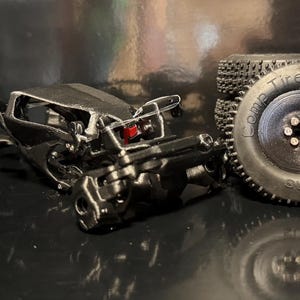 Puede incluir: Un chasis de crawler RC impreso en 3D de color negro con varillajes mecánicos expuestos y un pequeño componente rojo. Junto al chasis hay un neumático de goma negro con el texto Comp Tires 1.0 inch Mud Terrain grabado en el lateral y una rueda.