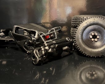 May include: A black 3D printed RC crawler chassis frame with exposed mechanical linkages and a small red component. Next to the frame is a black rubber tire with the text Comp Tires 1.0 inch Mud Terrain embossed on the side wall and a six-bolt wheel.