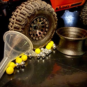 May include: A clear plastic funnel, a collection of small yellow and silver spherical weights, and a black textured wheel rim positioned in front of a large off-road tire with a deep tread pattern and a metallic center hub with visible bolts.