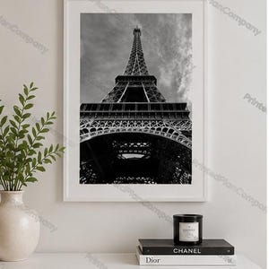 May include: A black and white framed wall art print featuring a low-angle perspective of the Eiffel Tower against a cloudy sky. The print is displayed in a white frame, positioned above a white surface with a candle and two stacked books.