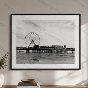 May include: A black and white framed wall art print featuring a Ferris wheel on a pier extending over a beach. The image is enclosed in a thin black frame with a white mat border, displaying a coastal scene with a cloudy sky and calm water.