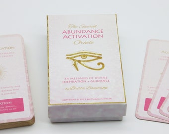 May include: A white box with gold trim and the text "The Sacred Abundance Activation Oracle" and "44 Messages of Divine Inspiration + Guidance by Britta Baumann". The box has a pink lid and a gold eye symbol on the front. The box is sitting on a white surface with a stack of pink and gold cards.
