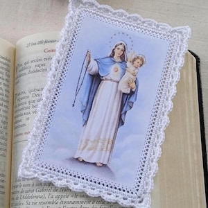 Peut inclure: Un signet religieux avec une bordure en dentelle crochetée blanche, représentant une image de la Vierge Marie tenant l'enfant Jésus. Le signet est posé sur un livre ouvert, avec du texte visible sur les pages.