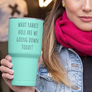 Könnte beinhalten: Ein türkisfarbener Isolierbecher mit dem Text "WHAT RABBIT HOLE ARE WE GOING DOWN TODAY?" in Schwarz. Der Becher wird von einer Person gehalten, die eine Jeansjacke und einen rosa Schal trägt.