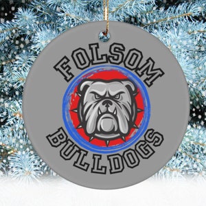 May include: A gray and white ceramic ornament with a red, white, and blue bulldog mascot. The ornament reads "Folsom Bulldogs" in a black font.