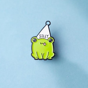 Puede incluir: Un pequeño pin de esmalte que muestra una rana de color verde brillante con un gorro de fiesta blanco que tiene la palabra SILLY impresa en letras negras. La rana tiene ojos negros grandes y una pequeña lengua afuera, con un borde negro.