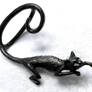 Puede incluir: Un pendiente de oreja en forma de gato gris oscuro. El gato está en una pose juguetona, con su pata delantera extendida. El pendiente se curva hacia arriba y alrededor de la oreja. El metal tiene un aspecto texturizado.
