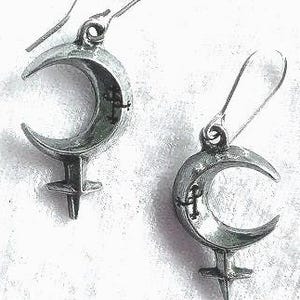 May include: A pair of silver-toned earrings. Each earring features a crescent moon shape with a cross symbol inside, and a stylized female symbol below. The earrings have a hook closure.