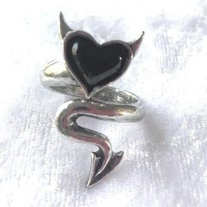 Puede incluir: Un anillo plateado con una piedra negra en forma de corazón, cuernos de diablo y un diseño de cola. El anillo tiene un estilo retorcido y envolvente, creando una estética única y atrevida. El anillo es probablemente de metal.