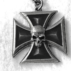 May include: A silver-coloured pendant necklace featuring a skull centered on a Maltese cross design. The cross has a beveled edge and a dark, textured finish. The chain is a simple, linked design.