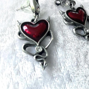 Puede incluir: Pendientes plateados con diseño de corazón de diablo. Cada pendiente tiene un centro en forma de corazón rojo, pequeños cuernos y un remolino en forma de cola con un acento de cristal transparente. Sobre una superficie blanca texturizada.