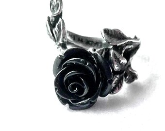 Könnte beinhalten: Ein silberfarbener Ring mit einer markanten, dunkel mattschwarzen Rose als Mittelstück. Die Ringschiene ist mit geformten metallischen Blättern und Stielen gestaltet, die sich um den Finger legen und ein detailliertes Blumenmotiv bilden.