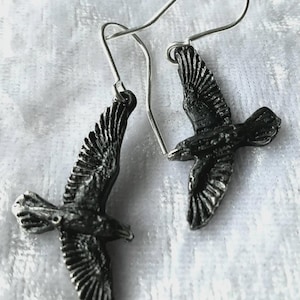 Puede incluir: Un par de pendientes plateados con un diseño detallado de un pájaro en vuelo. Los pendientes tienen un cierre de gancho y están colocados sobre un fondo blanco texturizado. Las alas del pájaro están extendidas, mostrando detalles intrincados de plumas.