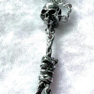 Puede incluir: Collar con colgante plateado que presenta un dije de calavera y un diseño de soga corrediza. La calavera tiene cuencas de los ojos y una superficie texturizada. La soga corrediza está intrincadamente elaborada con detalles de cuerda trenzada.