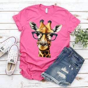 May include: Pink t-shirt with a graphic of a giraffe wearing glasses. The giraffe is brown and yellow with a black nose and black eyes. The glasses are black.