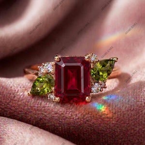May include: A rose gold ring featuring a large emerald-cut deep red center stone, flanked by two pear-cut olive green stones and accented with small round clear stones on each side, set in a polished metal band with a warm metallic finish.