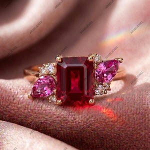Peut inclure: Une bague en or avec une pierre précieuse centrale rouge foncé taille émeraude, flanquée de deux pierres roses taille poire et de petites pierres d'accent rondes et claires. L'anneau est en or poli et le sertissage forme un groupe.