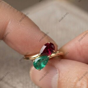 May include: A gold ring featuring two pear-shaped gemstones, one deep red ruby and one vibrant green emerald, set in a bypass style band. The stones are positioned side-by-side, creating a contrasting colour display on a polished gold metal band.
