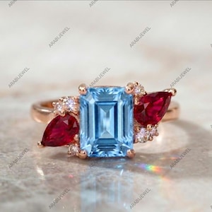 May include: A rose gold ring featuring a central emerald-cut light blue gemstone flanked by two pear-cut deep red gemstones. The setting is accented with small, round, clear white stones clustered around the larger colored gems.