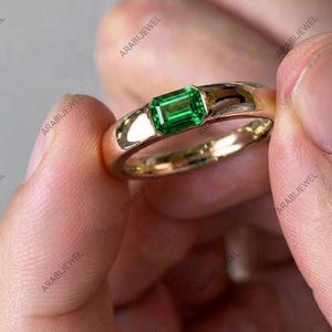 May include: A gold ring featuring a single emerald-cut green gemstone set in a smooth, polished metal band. The ring is held between two fingers, showcasing the vibrant green stone and the reflective, warm-toned gold surface of the band.
