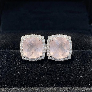 Puede incluir: Un par de pendientes de botón con gemas de cuarzo rosa pálido en talla cojín. Cada piedra está rodeada por un halo de pequeñas piedras blancas redondas engastadas en un marco metálico. El texto ARABIJEWEL es visible en cada gema.