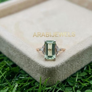 May include: A ring featuring a large, rectangular, light green emerald-cut gemstone set in a rose gold band. The center stone is flanked by small, clear baguette and round-cut accent stones. The text ARABIJEWELS is visible above the ring.