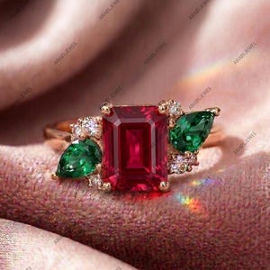 May include: A gold ring featuring a large emerald-cut red center stone flanked by two pear-cut green stones and small round clear accent stones. The band is polished gold, and the setting holds the stones in a cluster arrangement on the ring.