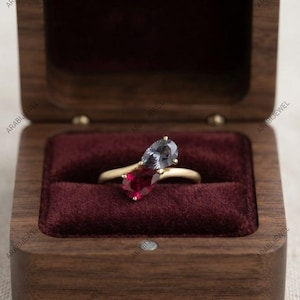 Pode incluir: Um anel de ouro com duas pedras preciosas em corte de pera, uma vermelho-profundo e outra azul-acinzentada, montadas em estilo bypass. O anel é exibido dentro de uma caixa de madeira escura com um forro de veludo bordô que contrasta com as pedras.