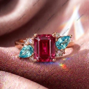 May include: A gold ring featuring a large emerald-cut red gemstone in the center, flanked by two pear-cut light blue gemstones. Small, clear, round-cut accent stones are clustered around the base of the larger stones on the gold band.