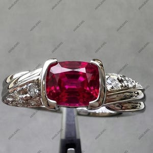 May include: A silver-toned ring featuring a central cushion-cut vibrant red gemstone held in a tension setting. The band is crafted with a polished finish and includes small, clear, round-cut stones embedded along the sides of the shank.