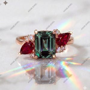 May include: A rose gold ring featuring a central emerald-cut dark green gemstone flanked by two pear-cut deep red rubies. The setting includes small round clear accent stones on both sides of the center stone, creating a multi-gemstone design.