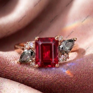 May include: A rose gold ring featuring a large emerald-cut red center stone flanked by two pear-cut grey side stones. The setting is accented with small round clear stones clustered around the base of the larger gems on a polished metal band.