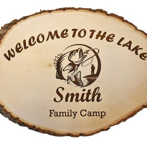 May include: Oval wooden sign with a natural bark edge. The sign reads "WELCOME TO THE LAKE" and features a fishing-themed graphic with the name "Smith" and "Family Camp" below. The wood has a light, natural finish.