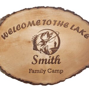 May include: Oval wooden sign with a natural bark edge. The sign reads "WELCOME TO THE LAKE" and features an engraved image of a fish jumping out of the water with a fisherman. The name "Smith" and "Family Camp" are also engraved.