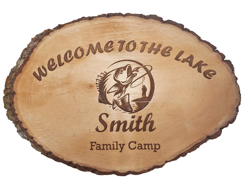 May include: Oval wooden sign with a natural bark edge. The sign reads "WELCOME TO THE LAKE" and features an engraved image of a fish jumping out of the water with a fisherman. The name "Smith" and "Family Camp" are also engraved.