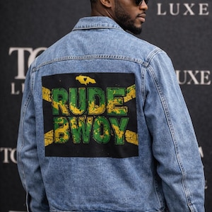 Puede incluir: Chaqueta vaquera azul claro con un parche negro con las palabras "RUDE BWOY" en verde y amarillo, con un diseño de bandera jamaicana. La chaqueta tiene un diseño clásico con botones.