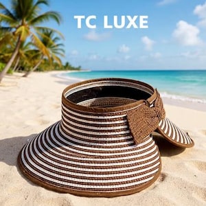 May include: A wide-brimmed sun visor with brown and white stripes, a brown bow, and the text "TC LUXE" in white. The visor is on a sandy beach with palm trees and the ocean in the background.