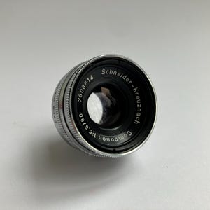 Puede incluir: Un objetivo Schneider-Kreuznach plateado y negro con el texto "Componon 1:5.6/80" y "7808814" impreso. El objetivo es cil&iacute;ndrico con un anillo de enfoque estriado.