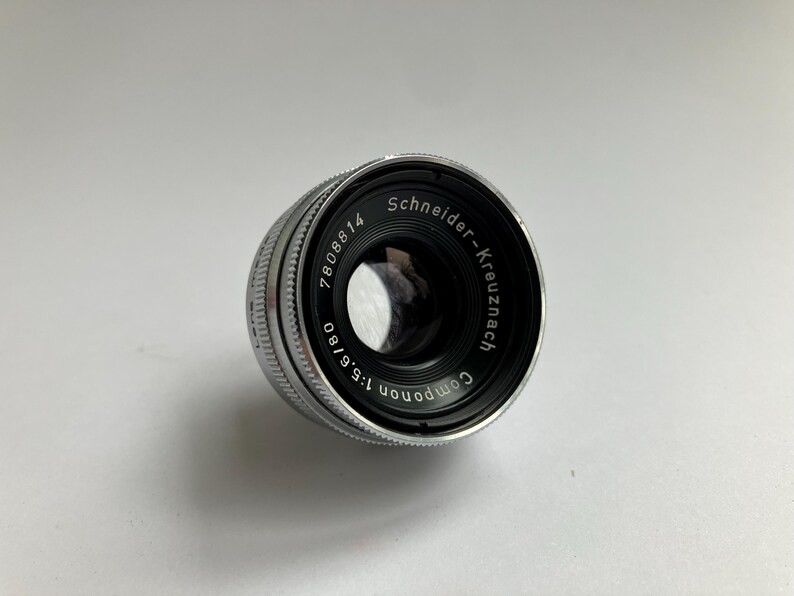 Puede incluir: Un objetivo Schneider-Kreuznach plateado y negro con el texto "Componon 1:5.6/80" y "7808814" impreso. El objetivo es cil&iacute;ndrico con un anillo de enfoque estriado.